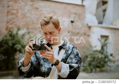 Young Caucasian man adjusting vintage camera outdoors, preparing for shot in urban environment with greenery and rustic walls behind Young Caucasian man adjusting vintage camera outdoors, preparing for shot in urban environment with greenery and rustic walls behind 118038572