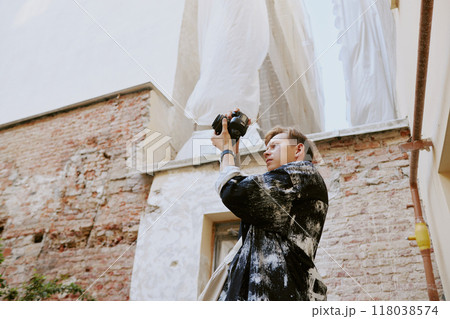 Man operating a handheld gadget while capturing images of tall buildings. Puffy white clouds in the background with rustic brick buildings surrounding him 118038574