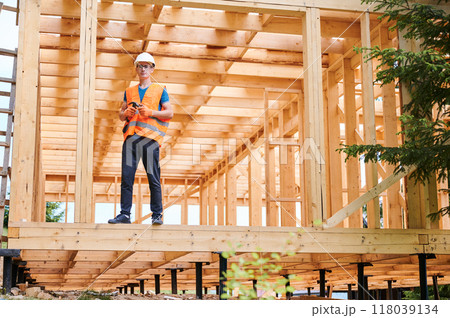 Carpenter constructing wooden frame two-story house near the forest. Man in glasses holding hammer, dressed in protective helmet and orange safety vest. Concept of ecological modern construction. 118039134