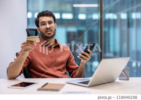 Frustrated man holding bank card and phone at office desk with laptop expressing concern about online fraud. Concept of financial problem, fraud, and technology misuse 118040286