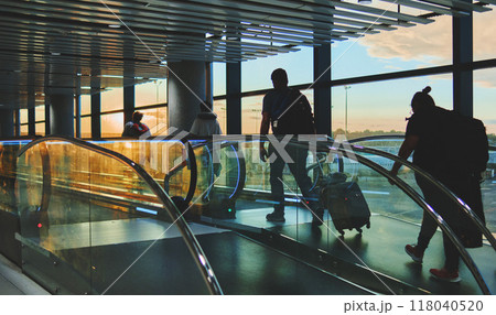 View on the aiport window with woman walking with suitcase at the departure hall during the sunset. Wide angle view with copy space. View on the aiport window with woman walking with suitcase at the departure hall during the sunset. Wide angle view with copy space. 118040520