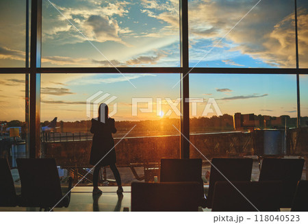 View on the aiport window with woman walking with suitcase at the departure hall during the sunset. Wide angle view with copy space. View on the aiport window with woman walking with suitcase at the departure hall during the sunset. Wide angle view with copy space. 118040523
