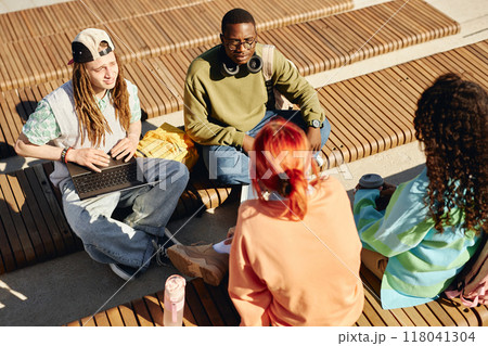 Diverse group of male students using laptop chatting with teenage girls while sitting on benches squinting in midday sun during friend gathering after classes Diverse group of male students using laptop chatting with teenage girls while sitting on benches squinting in midday sun during friend gathering after classes 118041304