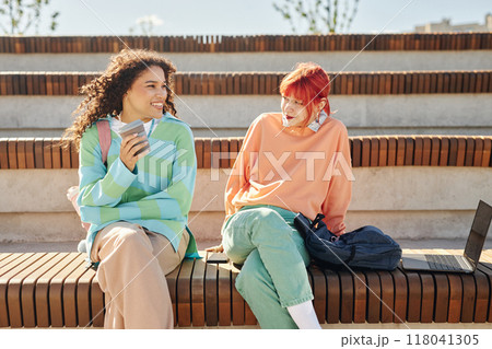 Smiling multiethnic girl hanging out with teenage female friend both dressed in pastel clothes while enjoying communication and sunny day on amphitheater seat at park 118041305