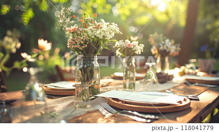 In a sunlit garden, a beautifully arranged table displays rustic plates and glasses, surrounded by elegant jars overflowing with vibrant flowers, creating a warm, inviting atmosphere 118041779