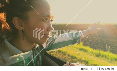 Female traveler smiles sticking hand out of car window, catches air currents, enjoying freedom, trip by car. Happy satisfied woman traveling by car, enjoying vacation trip, leaning out of car window. 118042616