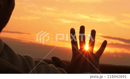 Silhouette of hand at orange sunset. Man stretches hand towards sun, warming palm in summer rays. Unity with nature, enjoyment of trip. Guy gains strength, contemplating, enjoying solitude with nature Silhouette of hand at orange sunset. Man stretches hand towards sun, warming palm in summer rays. Unity with nature, enjoyment of trip. Guy gains strength, contemplating, enjoying solitude with nature 118042691