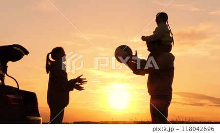Family playing with ball in nature at sunset, silhouettes. Daughter sits on father's shoulders, throwing ball with mother. Family travel adventures by car, active leisure. Weekend road trip to nature. Family playing with ball in nature at sunset, silhouettes. Daughter sits on father's shoulders, throwing ball with mother. Family travel adventures by car, active leisure. Weekend road trip to nature. 118042696