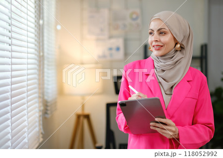 A woman in a pink jacket is holding a tablet and writing on a notepad A woman in a pink jacket is holding a tablet and writing on a notepad 118052992