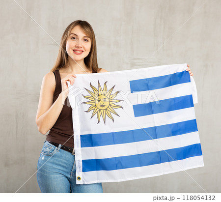 Happy cheerful young woman waving national flag of Uruguay while looking at camera at gray wall background 118054132