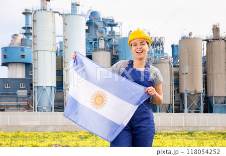 Young female engineer in helmet waving state flag of Argentina while standing in front of big tanks at chemical plant 118054252