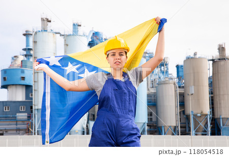 Upset disappointed young female engineer in helmet waving state flag of Bosnia and Herzegovina during strike in front of big tanks at chemical plant 118054518