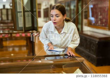 Attentive girl visitor at museum looks at exposition behind glass display case 118055368