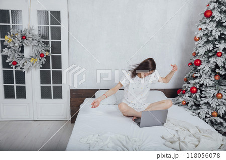Woman in pajamas with laptop on bed in bedroom Christmas tree with gifts 118056078