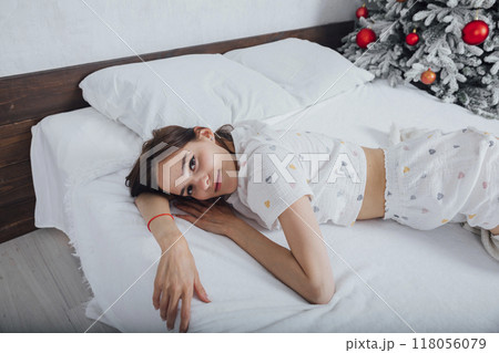 Woman in pajamas on bed in bedroom woke up by Christmas tree with gifts 118056079