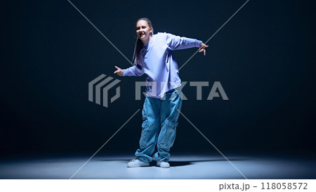 Youthful energy. Dynamic image of young girl, hip hop dancer performing on stage with passion against navy blue studio background Youthful energy. Dynamic image of young girl, hip hop dancer performing on stage with passion against navy blue studio background 118058572