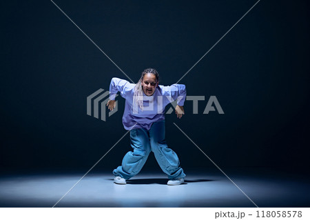 Dynamic image of young girl, passionate dancer in oversized clothes dancing hip hop with energy against navy blue studio background 118058578