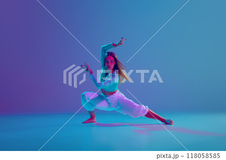 Dance club member. Young talented and expressive girl, dancer making energetic hip hop performance against gradient blue background in neon light Dance club member. Young talented and expressive girl, dancer making energetic hip hop performance against gradient blue background in neon light 118058585