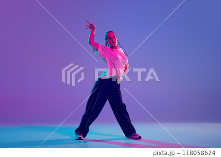 Young beautiful girl in stylish clothes performing hip hop with passion and energy against gradient purple background in neon light Young beautiful girl in stylish clothes performing hip hop with passion and energy against gradient purple background in neon light 118058624
