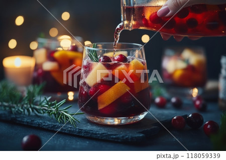Female Hand Pouring Holiday Sangria in Glasses with Fruit Slice, Cranberry, and Rosemary on Dark Christmas Background 118059339