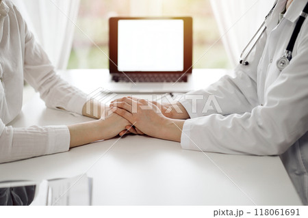 Doctor and patient sitting at the white table near window in clinic. Female physician's hands reassuring woman. Medicine concept Doctor and patient sitting at the white table near window in clinic. Female physician's hands reassuring woman. Medicine concept 118061691