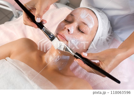 Professional beautician is applying mask to woman's face using two brushes. 118063596