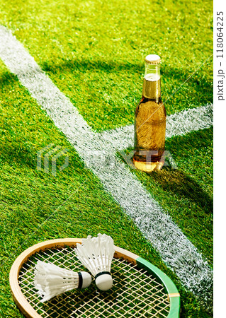 Badminton racket and shuttlecocks rest on green turf beside cold beer bottle. Perfect blend of sport and relaxation on sunny day, captured on field. 118064225
