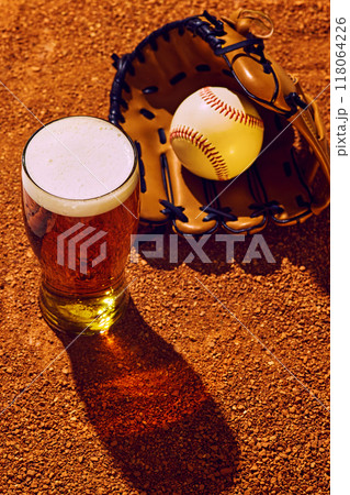Cold pint of beer rests on baseball field next to glove and ball, capturing perfect mix of game day excitement and refreshing break under sun. 118064226