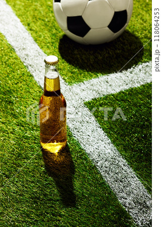 Glass beer bottle stands upright on lush green field. White line crosses stadium diagonally behind bottle, casting shadow on field. Glass beer bottle stands upright on lush green field. White line crosses stadium diagonally behind bottle, casting shadow on field. 118064253