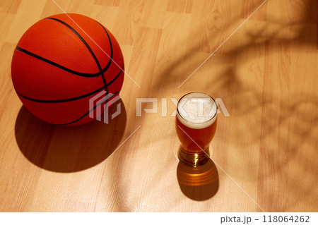 Glass of foamy, golden beer sits on wooden floor. Sunlight casts shadow of basketball hoop on court, floor, while basketball rests nearby. 118064262