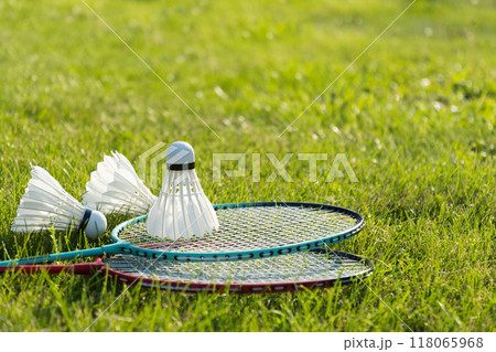 White badminton shuttlecock and badminton rackets on green grass in sunny shadow. Summer. Leisure games. Outdoors. Close up. 118065968