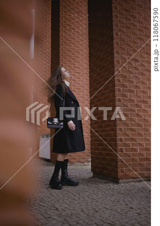 Young woman wearing a black coat poses near textured brick columns on a cloudy day 118067590