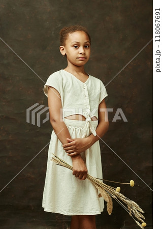 Vertical portrait of young calm African American girl in white dress holding bouquet of dried flowers while looking at camera standing on brown background in studio, copy space 118067691
