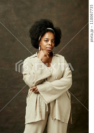 Vertical portrait shot of pensive African American woman with curly hair wearing beige casual clothes looking at camera while posing on brown background at studio 118067735