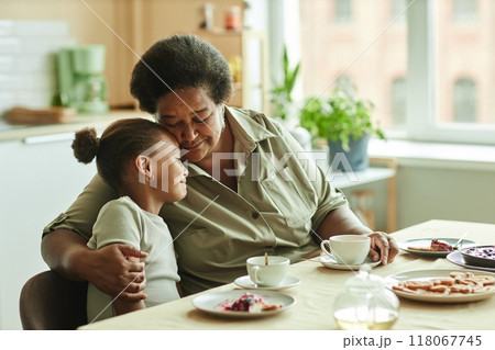 Medium shot of doting grandma gently embracing little granddaughter with love and affection while having tea sitting at table against window at sunlit kitchen, copy space 118067745