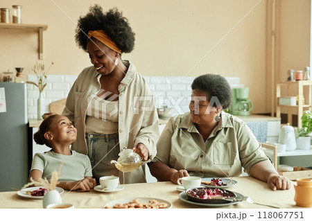 Smiling little girl with loving mother and granny having delicious cake sitting at table while mom pouring tea to daughters cup at beige kitchen 118067751