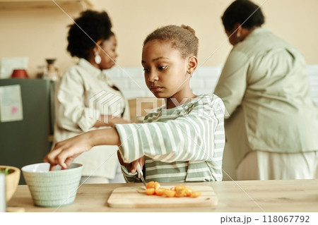 Medium shot of little Black girl in casual clothes taking vegetable from bowl cutting tomatoes while cooking fresh salad for healthy dinner at cozy beige kitchen, copy space 118067792