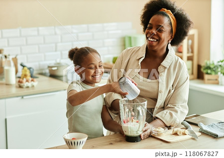 Laughing African American young mother and little daughter enjoying quality time while cooking together making fruit smoothie in blender at beige kitchen, copy space 118067827