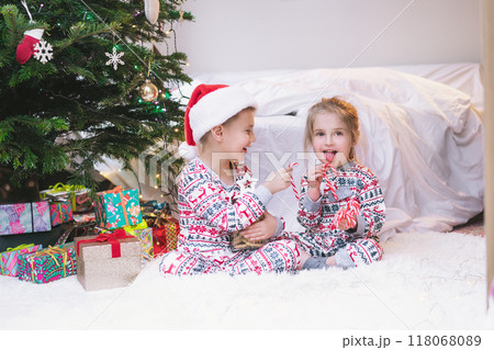 Children in festive pajamas eating Christmas candy canes from jar of holiday treats beside decorated Christmas tree during the festive season. Joyful siblings sit on cozy rug, enjoying festive sweets Children in festive pajamas eating Christmas candy canes from jar of holiday treats beside decorated Christmas tree during the festive season. Joyful siblings sit on cozy rug, enjoying festive sweets 118068089