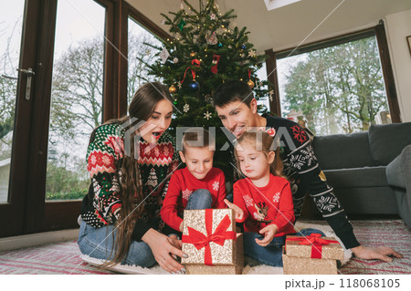 Happy family in festive sweaters sitting under Christmas tree at home. Emotional parents and kids feeling excited unwrapping Christmas gifts, having fun, Family time, childhood miracle time concept. Happy family in festive sweaters sitting under Christmas tree at home. Emotional parents and kids feeling excited unwrapping Christmas gifts, having fun, Family time, childhood miracle time concept. 118068105