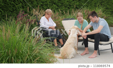 Family members, including a senior woman in a wheelchair, enjoy a relaxed moment with their Golden Retriever outdoors in a serene garden. 118069574