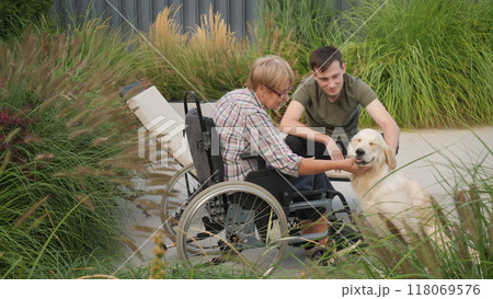 A senior woman in a wheelchair and a young man pet their Golden Retriever outdoors, sharing a joyful moment in a lush garden. 118069576