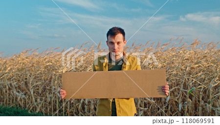A man holding a blank cardboard sign stands in a dry cornfield, wearing a yellow shirt against a blue sky. 118069591