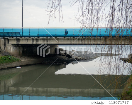 A man walks along the bridge along the seashore. Walking in the resort in the winter season. The man in the hood. Coast of the Black Sea. 118071757