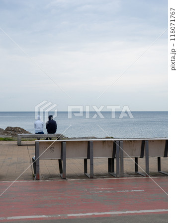 A couple is sitting on a bench by the sea. Cloudy weather at the resort. Not the season for tourism. Recreation of people on the coast. A couple is sitting on a bench by the sea. Cloudy weather at the resort. Not the season for tourism. Recreation of people on the coast. 118071767