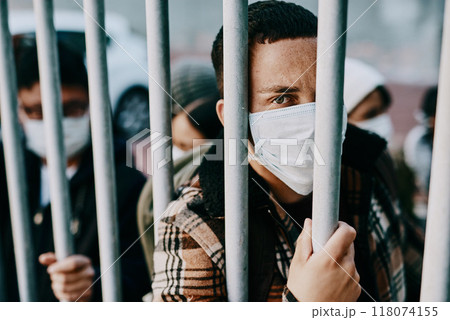 Who will make it out alive. Shot of a young man wearing a mask while stuck behind a gate in a foreign city. Who will make it out alive. Shot of a young man wearing a mask while stuck behind a gate in a foreign city. 118074155