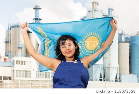 Positive girl in overalls with the Kazakhstan flag in her hands against the background of chimneys of modern factory Positive girl in overalls with the Kazakhstan flag in her hands against the background of chimneys of modern factory 118078359