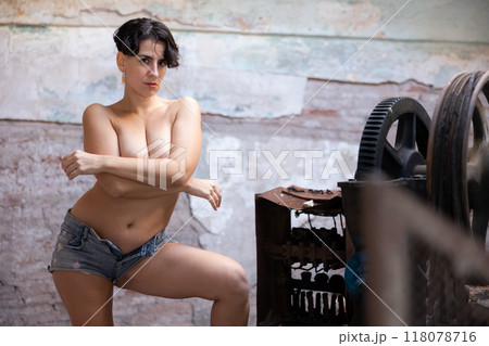 Brunette in t-shirt and blue short standing with bare breasts near old rusty machine tools in abandoned plant 118078716