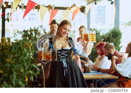 Beautiful smiling young woman in traditional Bavarian clothes, waitress holding mugs with lager beer, with group of people on background Beautiful smiling young woman in traditional Bavarian clothes, waitress holding mugs with lager beer, with group of people on background 118085754
