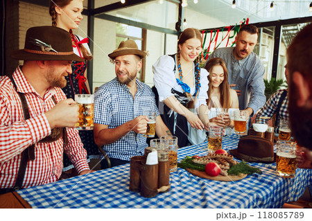 Oktoberfest joy. Men and women, friends sitting at table at local bar, enjoying beer. Celebration with Bavarian traditions Oktoberfest joy. Men and women, friends sitting at table at local bar, enjoying beer. Celebration with Bavarian traditions 118085789